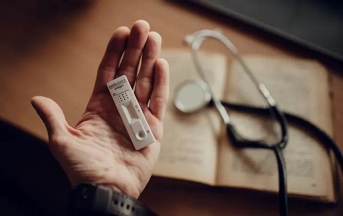 Hand holds a test device next to a stethoscope and an open book on a wooden desk during morning light
