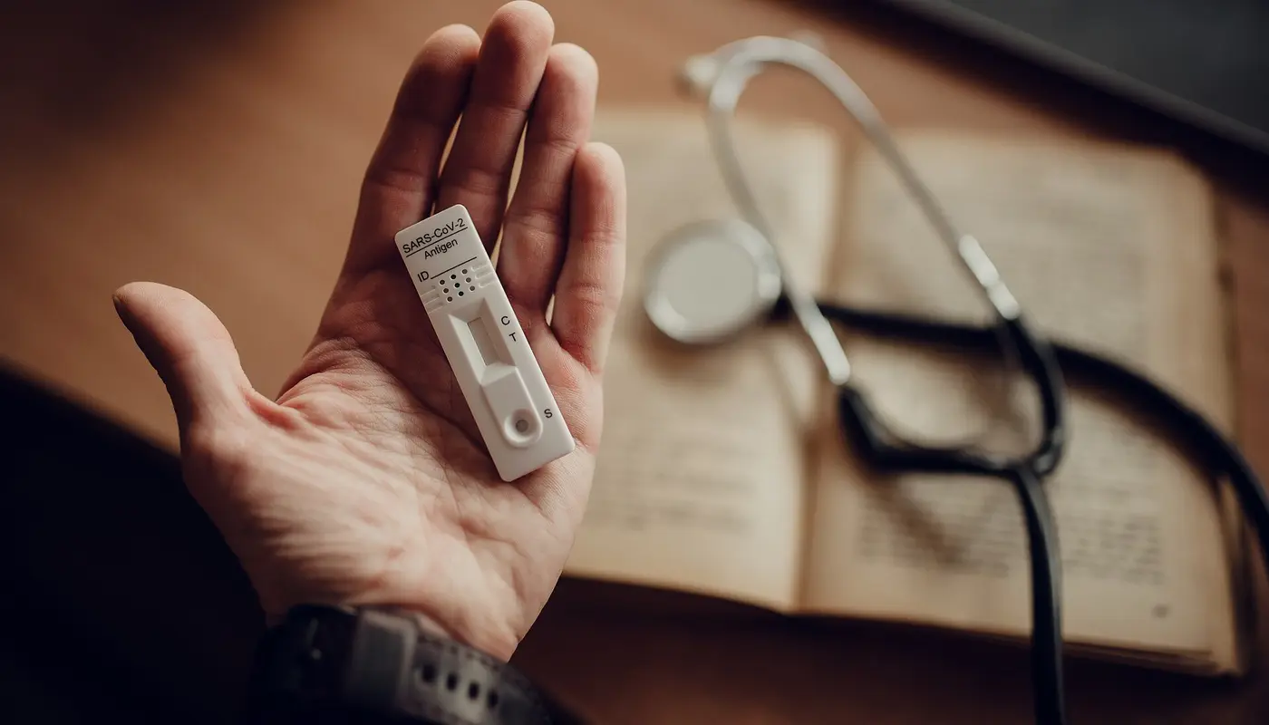 Hand holds a test device next to a stethoscope and an open book on a wooden desk during morning light